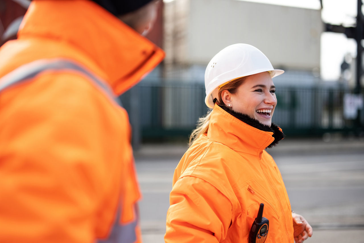 Female engineer smiling while talking with a worker at the shipyard. Two commercial dock workers in reflective uniform.