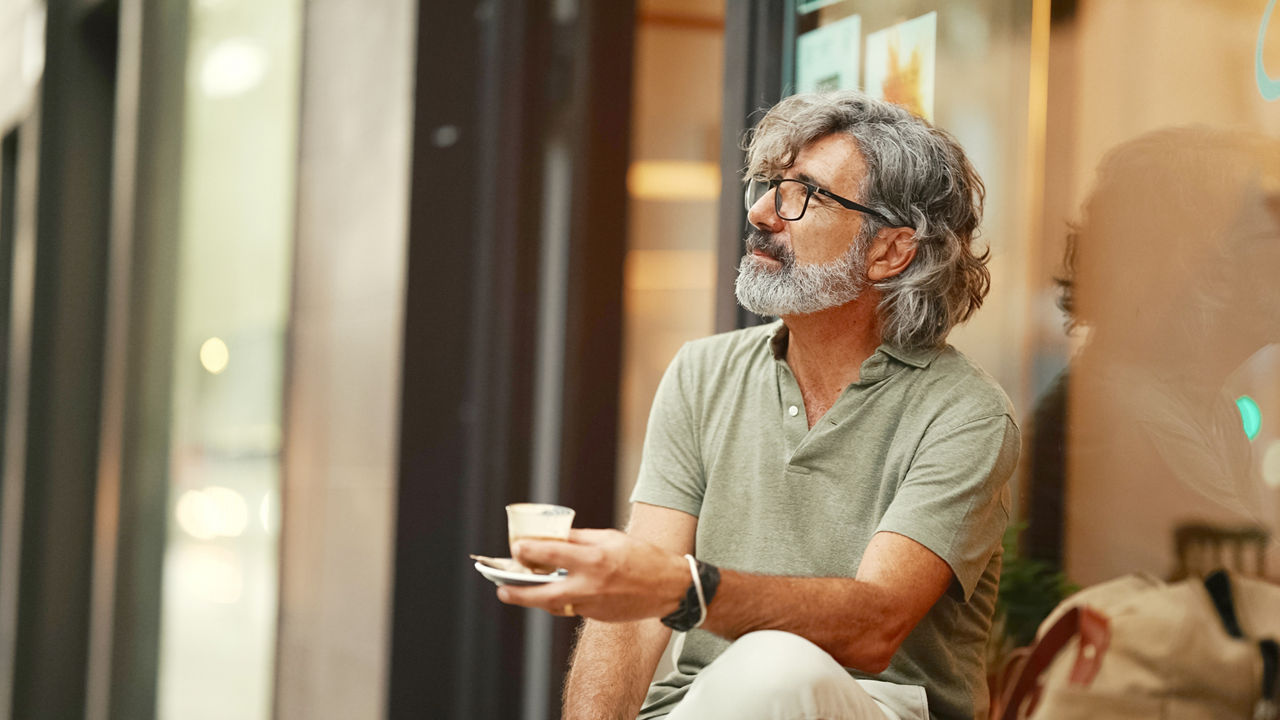 Clouse-up, thoughtful middle-aged man with gray hair and beard, wearing casual clothes, sits in street cafe. Mature gentleman in eyeglasses drinks aroma coffee
