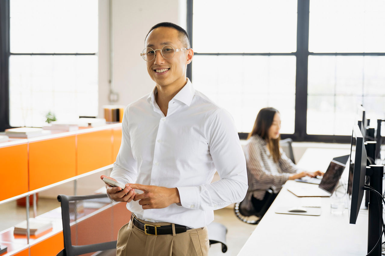 Young man with shaved head holding mobile phone in office, smiling towards camera