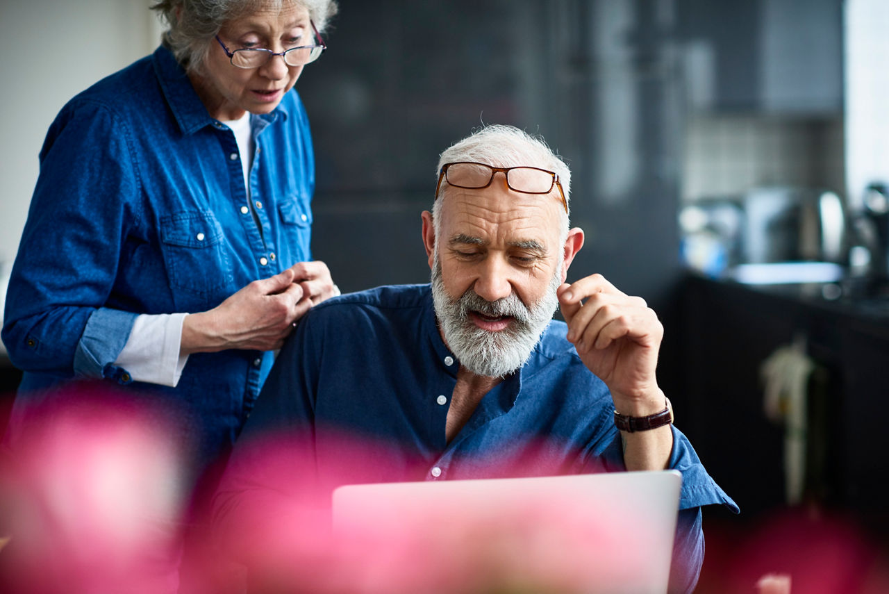 Candid portrait of senior couple at home, man with grey hair and beard working on computer, glasses resting on forehead, seniorpreneur working from home with wife