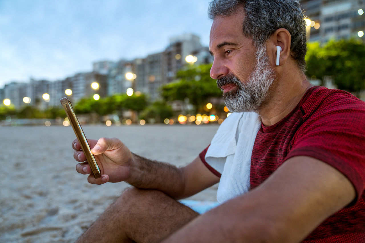 Close-up of an active senior man with a towel around his neck relaxing after training in the early hours of the morning before the sunrise, using his smartphone.