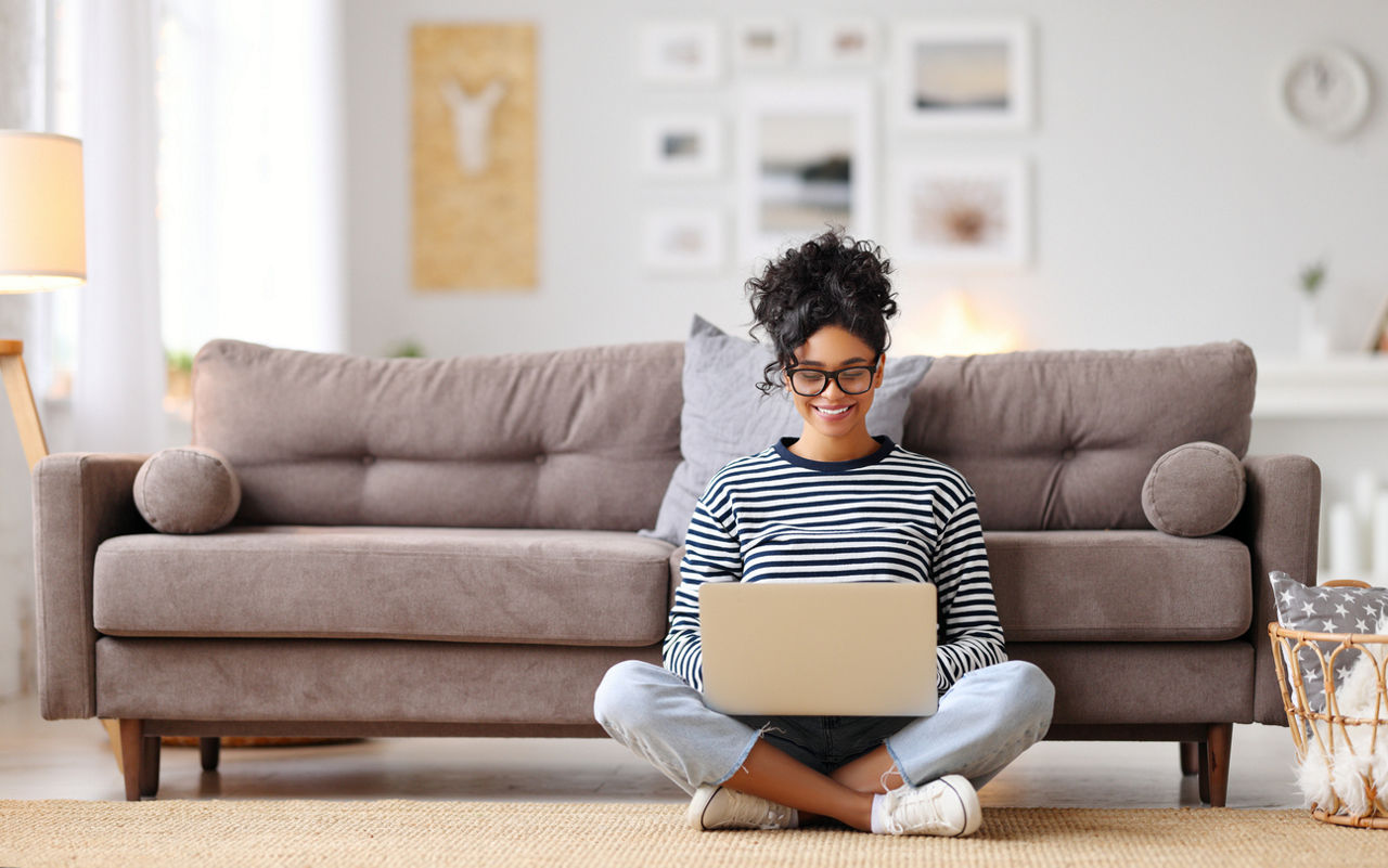 Cheerful young ethnic female freelancer in glasses and casual clothes focusing on screen and interacting with laptop while sitting alone on floor in light modern living room