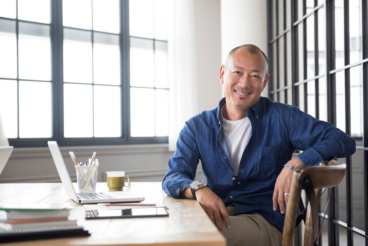 Portrait of mature Japanese smiling man sitting at desk with laptop computer and digital tablet.